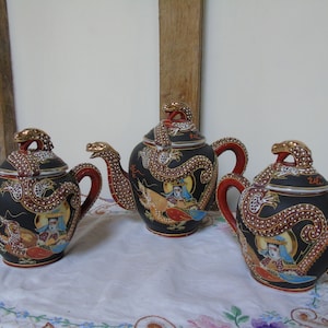 Op de afbeelding: Een set van drie zwarte en rode theepotten met gouden accenten en ingewikkelde drakenontwerpen. De theepotten zijn versierd met witte parel-achtige accenten en hebben kleurrijke figuren.