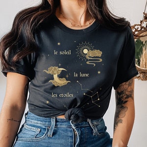 Puede incluir: Una camiseta negra con un dise&ntilde;o gr&aacute;fico dorado que presenta un sol, una luna y estrellas con el texto "le soleil", "la lune" y "les &eacute;toiles".