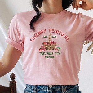 May include: A pink t-shirt with the text "CHERRY FESTIVAL 1926 1996 TRAVERSE CITY MICHIGAN" and a graphic of a basket of cherries.