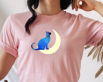 Op de afbeelding: Een roze T-shirt met een blauw katten silhouet dat op een gele halve maan zit. De kat kijkt naar rechts en de maan bevindt zich achter hem. Het shirt wordt gedragen door een persoon met denim shorts.