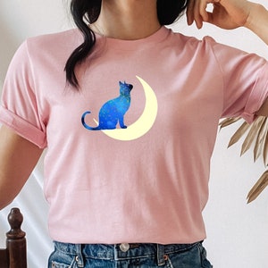 May include: A pink t-shirt with a blue cat silhouette sitting on a yellow crescent moon. The cat is facing to the right and the moon is behind it. The shirt is being worn by a person with denim shorts.