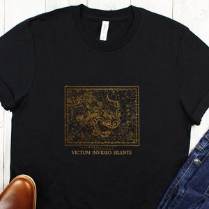 Puede incluir: Camiseta negra con un gráfico de constelación dorado y el texto "VICTUM INVIDIO SILENTE". La camiseta se combina con jeans azules y zapatos de cuero marrón.