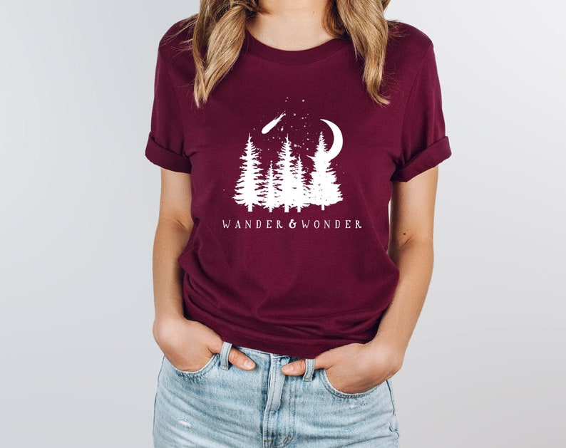 May include: A maroon t-shirt with a white graphic design of trees, a crescent moon, and a shooting star. The text "WANDER & WONDER" is printed below the graphic.