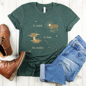 Puede incluir: Una camiseta verde oscuro con un dise&ntilde;o gr&aacute;fico dorado de un sol, una luna y estrellas. El texto en la camiseta dice "le soleil, la lune, les etoiles".