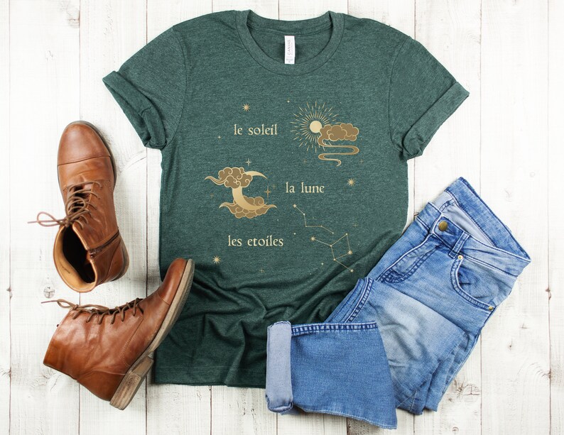 Puede incluir: Una camiseta verde oscuro con un dise&ntilde;o gr&aacute;fico dorado de un sol, una luna y estrellas. El texto en la camiseta dice "le soleil, la lune, les etoiles".
