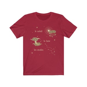 Puede incluir: Camiseta roja con un dise&ntilde;o gr&aacute;fico dorado de un sol, una luna y estrellas. El texto "le soleil", "la lune" y "les etoiles" est&aacute; escrito en franc&eacute;s.