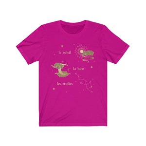 Puede incluir: Una camiseta rosa fucsia con un dise&ntilde;o gr&aacute;fico dorado de un sol, una luna y estrellas. El texto "le soleil", "la lune" y "les etoiles" est&aacute; impreso en dorado en la camiseta.