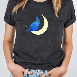 May include: A dark gray t-shirt with a blue cat silhouette sitting on a crescent moon. The cat and moon are filled with a galaxy-like pattern of blue and white.