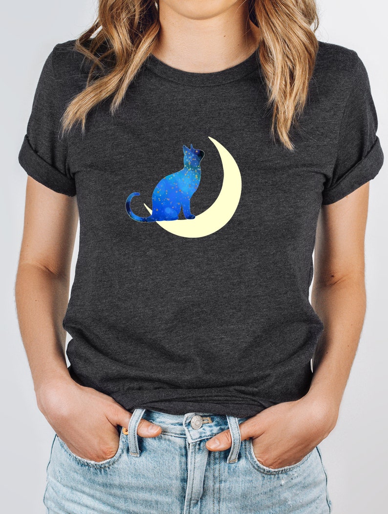 May include: A dark gray t-shirt with a blue cat silhouette sitting on a crescent moon. The cat and moon are filled with a galaxy-like pattern of blue and white.