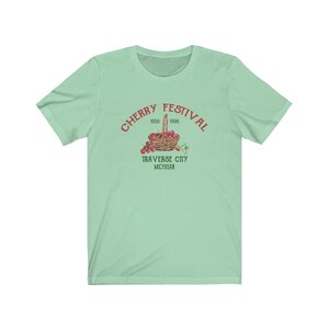 May include: A light green t-shirt with a red graphic of a basket of cherries. The graphic says "Cherry Festival 1926 1996 Traverse City Michigan".