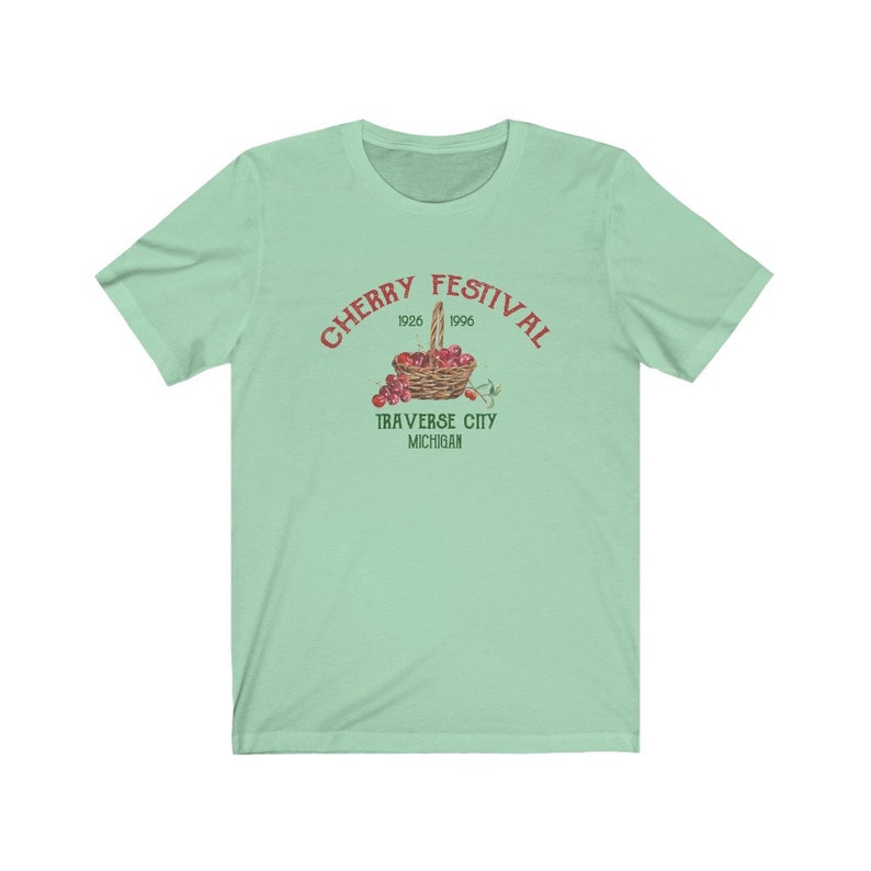 May include: A light green t-shirt with a red graphic of a basket of cherries. The graphic says "Cherry Festival 1926 1996 Traverse City Michigan".