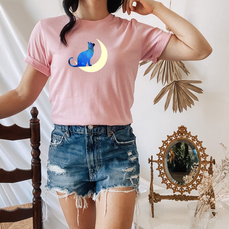 May include: A pink t-shirt with a blue cat silhouette sitting on a yellow crescent moon. The cat is facing to the right and the moon is behind it. The shirt is being worn by a person with denim shorts.