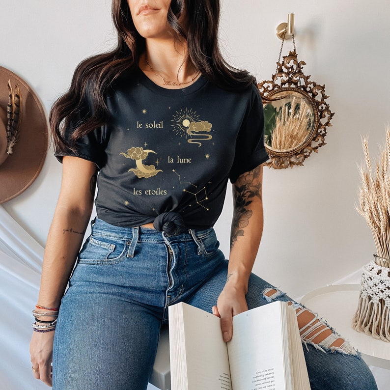 Puede incluir: Una camiseta negra con un dise&ntilde;o gr&aacute;fico dorado que presenta un sol, una luna y estrellas con el texto "le soleil", "la lune" y "les &eacute;toiles".