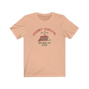 May include: A peach-colored t-shirt with a red graphic of a basket of cherries. The graphic says "Cherry Festival 1926 1996 Traverse City Michigan".
