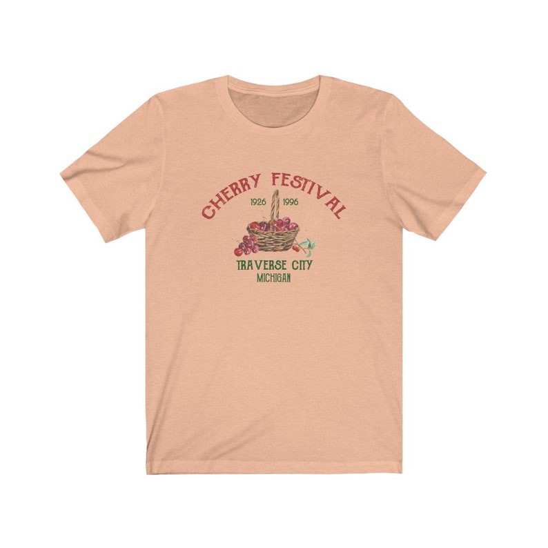 May include: A peach-colored t-shirt with a red graphic of a basket of cherries. The graphic says "Cherry Festival 1926 1996 Traverse City Michigan".