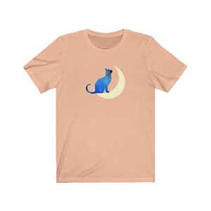 May include: A peach-colored t-shirt with a blue silhouette of a cat sitting on a crescent moon.