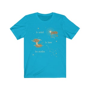Puede incluir: Una camiseta azul turquesa con un dise&ntilde;o gr&aacute;fico de un sol, una luna y estrellas. El texto en la camiseta dice "le soleil", "la lune" y "les &eacute;toiles".