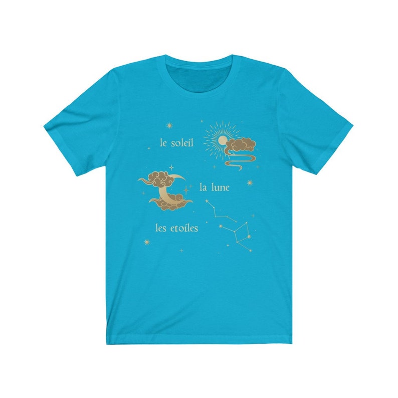 Puede incluir: Una camiseta azul turquesa con un dise&ntilde;o gr&aacute;fico de un sol, una luna y estrellas. El texto en la camiseta dice "le soleil", "la lune" y "les &eacute;toiles".