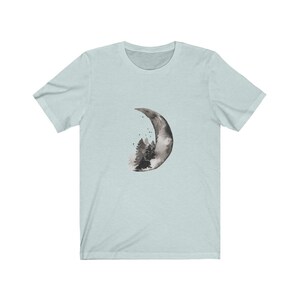 May include: A light blue t-shirt with a black and white watercolor design of a crescent moon with a forest inside.