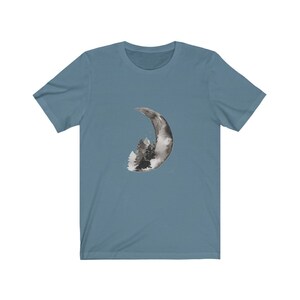 May include: A blue t-shirt with a black and white watercolor design of a crescent moon with a forest inside.