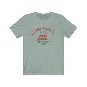 May include: A teal blue t-shirt with a red graphic of a basket of cherries. The graphic says "Cherry Festival 1926 1996 Traverse City Michigan".