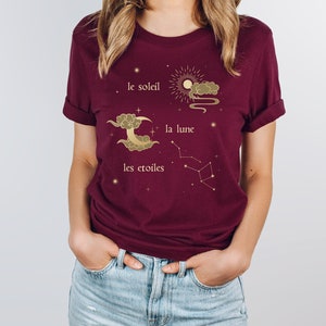 Puede incluir: Una camiseta color burdeos con un dise&ntilde;o gr&aacute;fico dorado que representa un sol, una luna y estrellas. El texto en la camiseta dice "le soleil", "la lune" y "les etoiles".