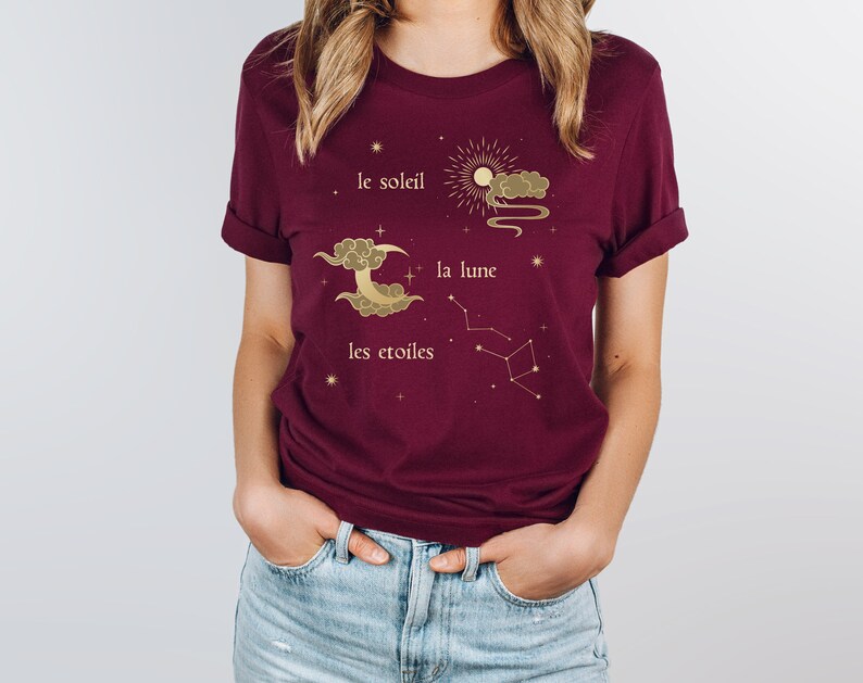 Puede incluir: Una camiseta color burdeos con un dise&ntilde;o gr&aacute;fico dorado que representa un sol, una luna y estrellas. El texto en la camiseta dice "le soleil", "la lune" y "les etoiles".