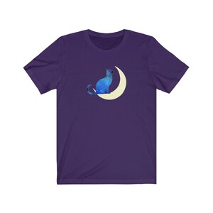May include: A purple t-shirt with a blue cat sitting on a crescent moon. The cat and moon are both outlined in white.