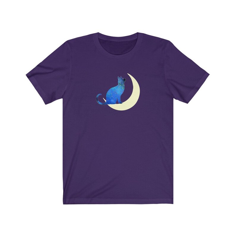 May include: A purple t-shirt with a blue cat sitting on a crescent moon. The cat and moon are both outlined in white.