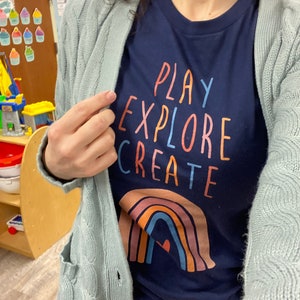 May include: A navy blue t-shirt with the words "PLAY EXPLORE CREATE" in a rainbow font. A rainbow with a heart in the center is below the text.