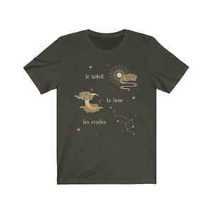 Puede incluir: Una camiseta verde oscuro con un dise&ntilde;o gr&aacute;fico dorado que presenta un sol, una luna y estrellas. El texto "le soleil", "la lune" y "les etoiles" est&aacute; impreso en dorado en la camiseta.