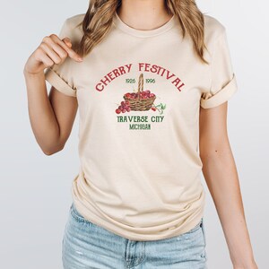 May include: A beige t-shirt with a red and green graphic that says "Cherry Festival 1926 1996 Traverse City Michigan" and a basket of red cherries.