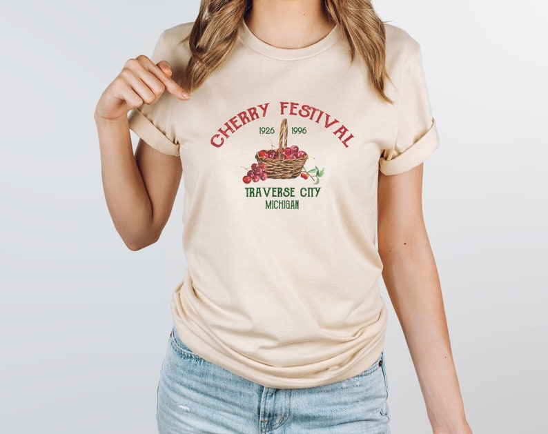 May include: A beige t-shirt with a red and green graphic that says "Cherry Festival 1926 1996 Traverse City Michigan" and a basket of red cherries.