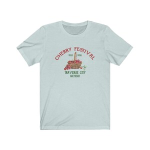 May include: A light blue t-shirt with a red and green graphic of a basket of cherries. The text "CHERRY FESTIVAL 1926 1996 TRAVERSE CITY MICHIGAN" is printed on the shirt.