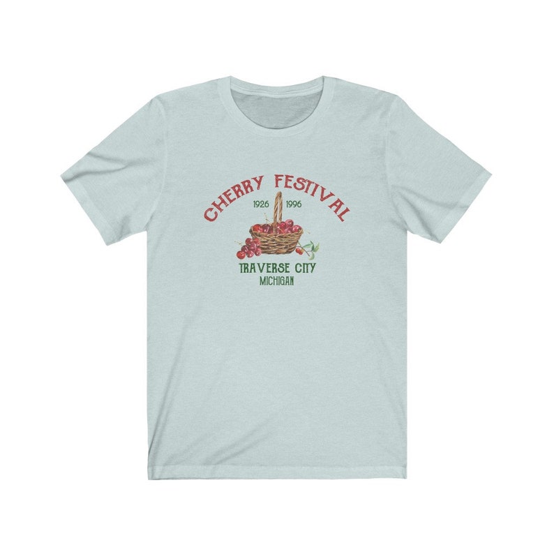 May include: A light blue t-shirt with a red and green graphic of a basket of cherries. The text "CHERRY FESTIVAL 1926 1996 TRAVERSE CITY MICHIGAN" is printed on the shirt.