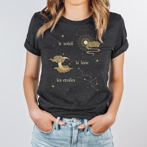 Puede incluir: Una camiseta gris oscuro con un dise&ntilde;o dorado que presenta un sol, una luna y estrellas. El texto en la camiseta dice "le soleil", "la lune" y "les &eacute;toiles".
