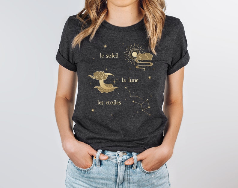 Puede incluir: Una camiseta gris oscuro con un dise&ntilde;o dorado que presenta un sol, una luna y estrellas. El texto en la camiseta dice "le soleil", "la lune" y "les &eacute;toiles".