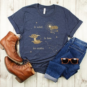 Puede incluir: Una camiseta azul marino con un gr&aacute;fico dorado de un sol, una luna y estrellas. El texto en la camiseta dice "le soleil", "la lune" y "les &eacute;toiles".