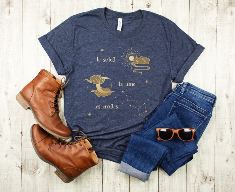 Puede incluir: Una camiseta azul marino con un gr&aacute;fico dorado de un sol, una luna y estrellas. El texto en la camiseta dice "le soleil", "la lune" y "les &eacute;toiles".