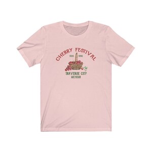 May include: Pink t-shirt with a vintage-style graphic of a basket of cherries. The text reads "Cherry Festival 1926 1996 Traverse City Michigan".