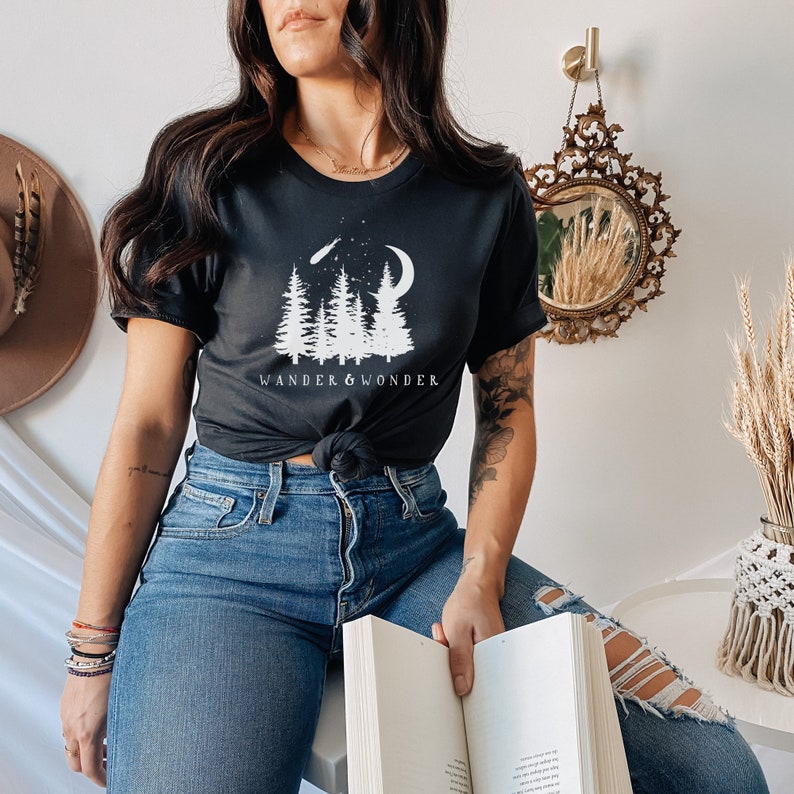 May include: A black t-shirt with a white graphic of trees, a crescent moon, and a shooting star. The text "WANDER & WONDER" is printed below the graphic.