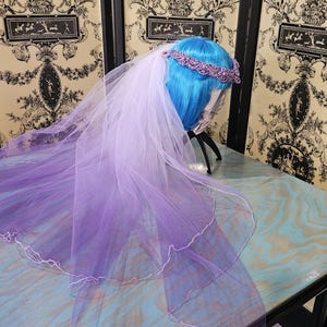 May include: A bridal veil with a purple ombre effect, transitioning from white to a deep violet. The veil is paired with a matching purple beaded headband. The veil is displayed on a mannequin head with blue hair.
