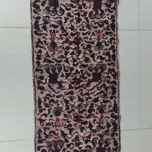 Può includere: Un pezzo di stoffa lungo e rettangolare con un motivo batik marrone e bianco. La stoffa ha un bordo rosso e marrone con un motivo ripetitivo.