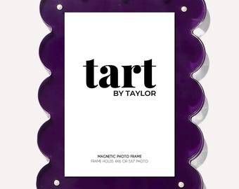 May include: A purple, scalloped-edge magnetic photo frame. The frame holds a 4x6 or 5x7 photo and features the text "tart by TAYLOR" in black. The frame has a glossy finish and is designed to hold a photo.
