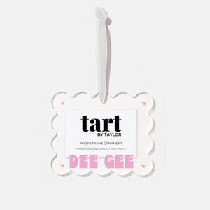 May include: A clear, scalloped-edge photo frame ornament with a white ribbon for hanging. The frame holds a 2x3 inch photo and features the text "tart by TAYLOR" and "DEE GEE" in black and pink.