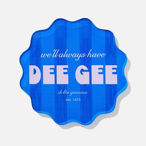 May include: A blue, scalloped-edge decorative item with vertical stripes. The text "we'll always have DEE GEE" is displayed in white, along with "delta gamma est. 1873". The item has a glossy finish and a slight shadow.