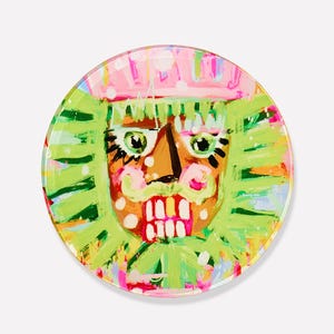May include: A round decorative plate featuring a colorful, whimsical illustration of a nutcracker face. The design includes a pink crown, green hair, and a brown face with exaggerated features. The plate has a glossy finish and a white background.