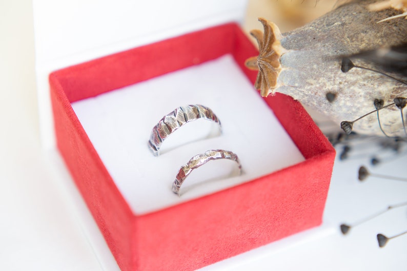 May include: Two silver wedding bands with a textured finish, displayed in a red velvet ring box.