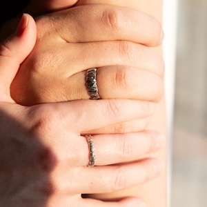 May include: Two silver rings on a person's fingers. The ring on the index finger has a textured, hammered design. The ring on the middle finger has a more delicate, textured design.