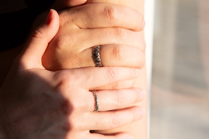 May include: Two silver rings on a person's fingers. The ring on the index finger has a textured, hammered design. The ring on the middle finger has a more delicate, textured design.
